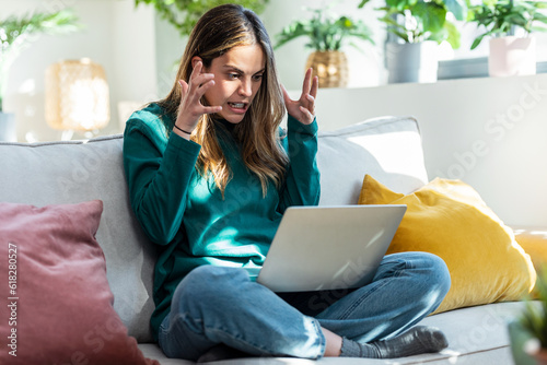 Angry frowning young woman working online on laptop sitting on couch at home