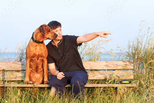 Young man sitting outdoors with his Rhodesian ridgeback dog on wooden bench and pointing finger at something ahead 