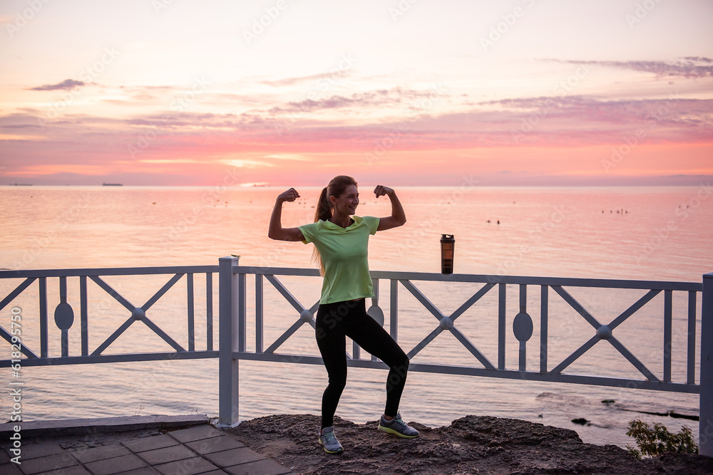 Fototapeta premium Young woman is fooling around in warm-up in morning at dawn of sun by sea. Sportswoman in sportswear