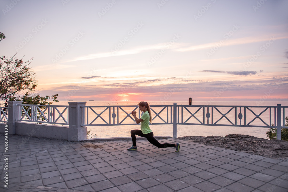 Young woman doing lunges, squats on warm-up in morning at sunrise by sea. Sportswoman in sportswear Young woman doing lunges, squats on warm-up in morning at sunrise by sea. Sportswoman in sportswear