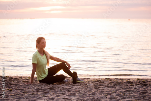 Young woman in sportswear is sitting on the seashore at sunrise. Girl is resting after workout