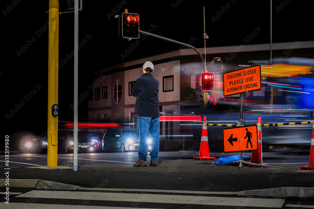 Man standing at traffic light. “Crossing Closed Please use Alternative Crossing” sign at intersection. Bus and car light trails on the road.