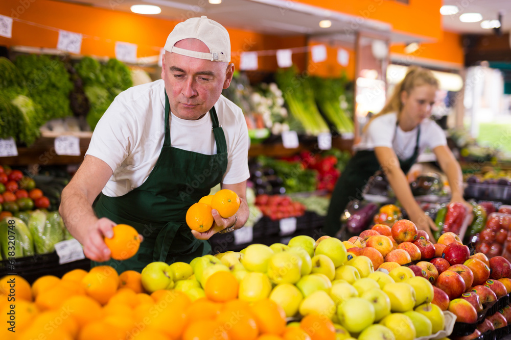 Fototapeta premium Focused man seller working in a supermarket in the vegetable department puts oranges on the counter for sale