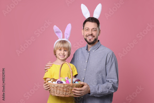 Father and son in bunny ears headbands with wicker basket of painted Easter eggs on pink background