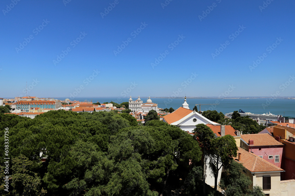 Uma deslumbrante vista aérea de Lisboa em um dia ensolarado, onde se mesclam áreas verdes, belas construções e o azul do mar ao fundo.