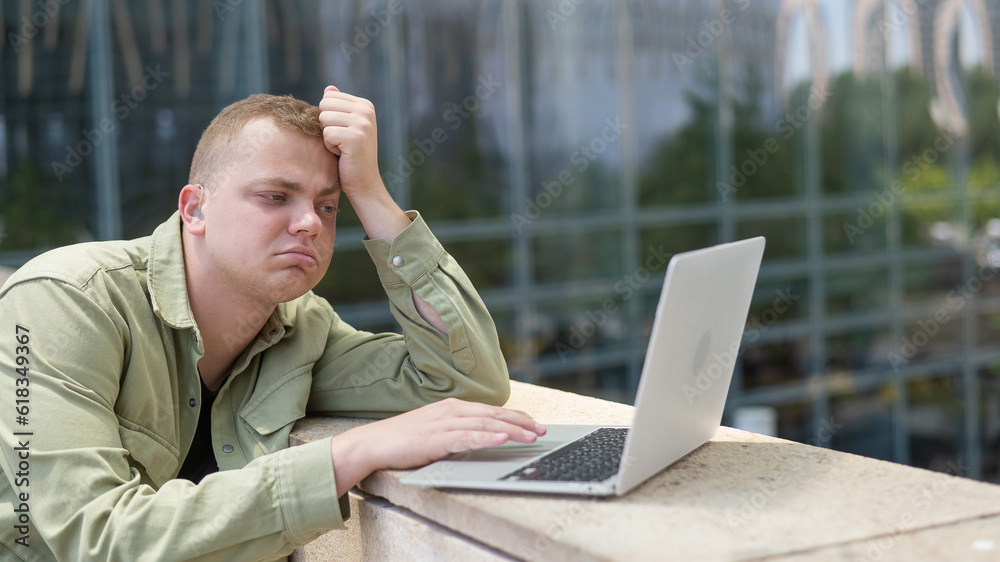 Sad guy with hearing aid using laptop outdoors. Sad guy with hearing aid using laptop outdoors.