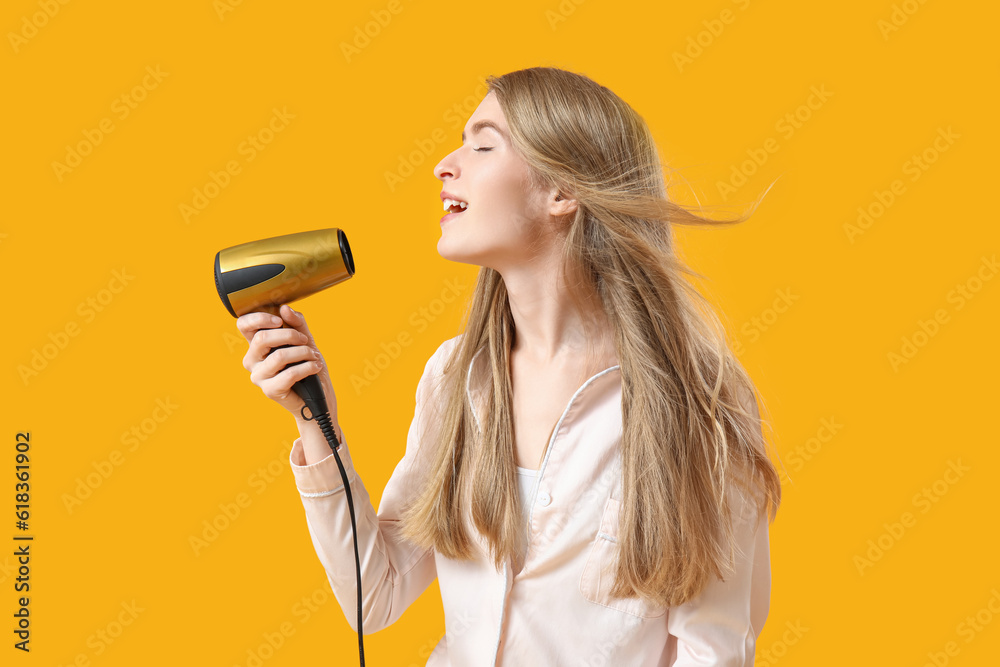 Young blonde woman in pajamas with hair dryer on yellow background Young blonde woman in pajamas with hair dryer on yellow background