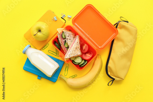 Different stationery, drink and lunch box with tasty food on yellow background