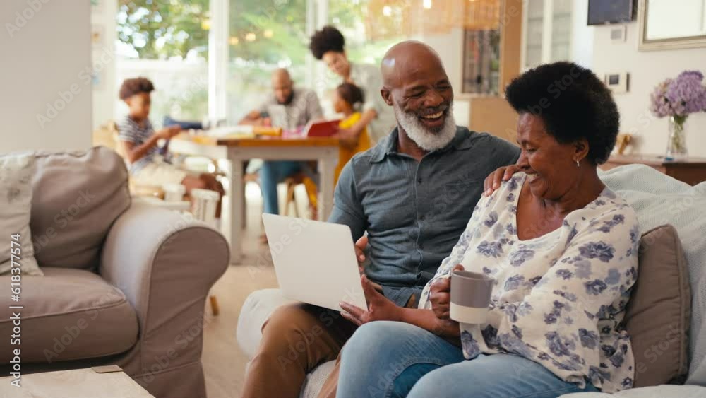 Senior couple at home looking at laptop together with multi-generation family in background - shot in slow motion