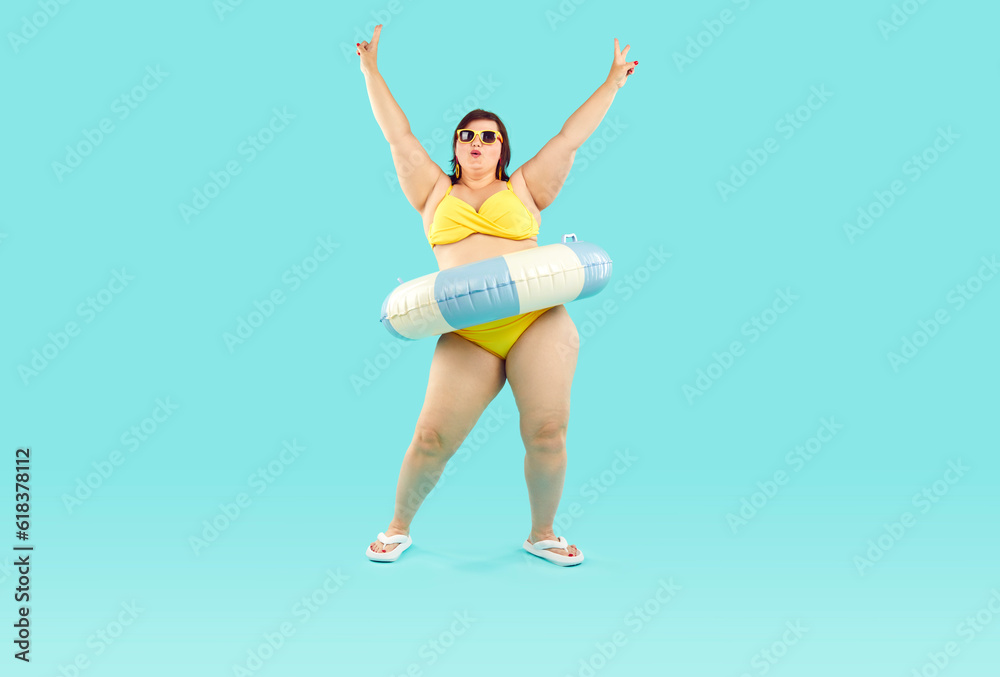 Happy confident fat woman having fun at beach party on summer vacation holiday. Plus size lady in yellow bikini, with inflatable rubber floater around waist dancing isolated on blue background