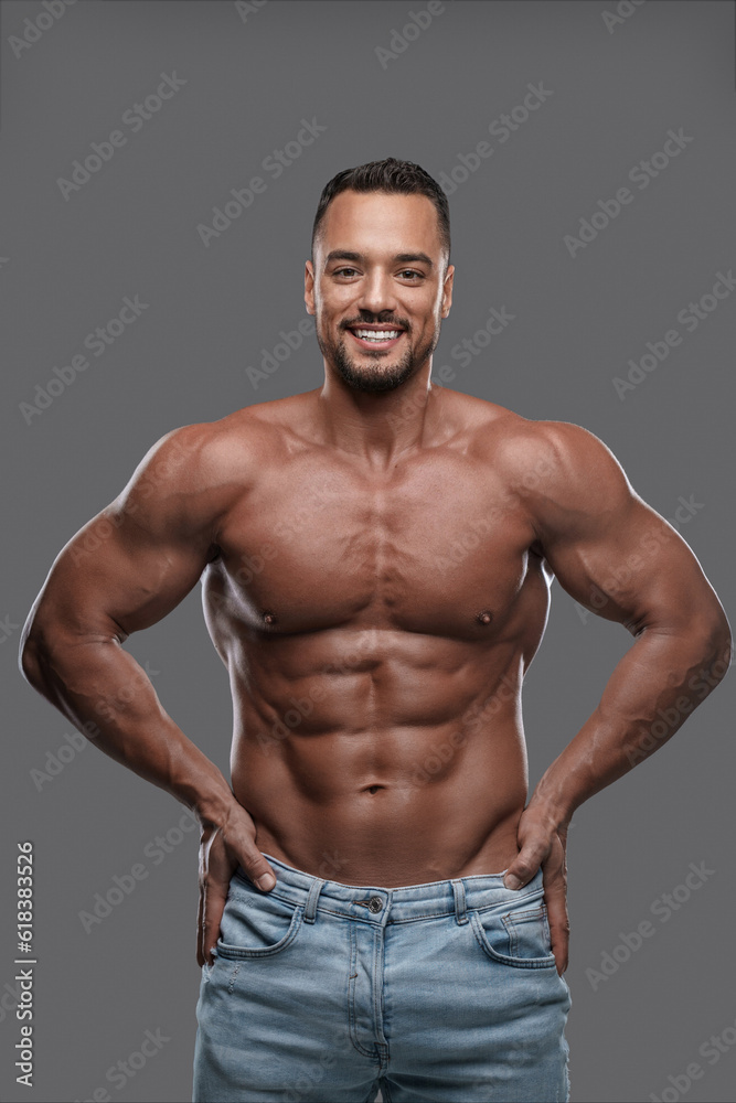 Charming and muscular male model with a dazzling smile poses shirtless against a neutral grey backdrop, emanating confidence and sex appeal