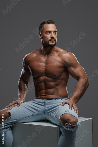 Remarkably attractive male model with a muscular physique poses topless in ripped jeans on a white cube against a grey backdrop, exuding sexiness and ruggedness