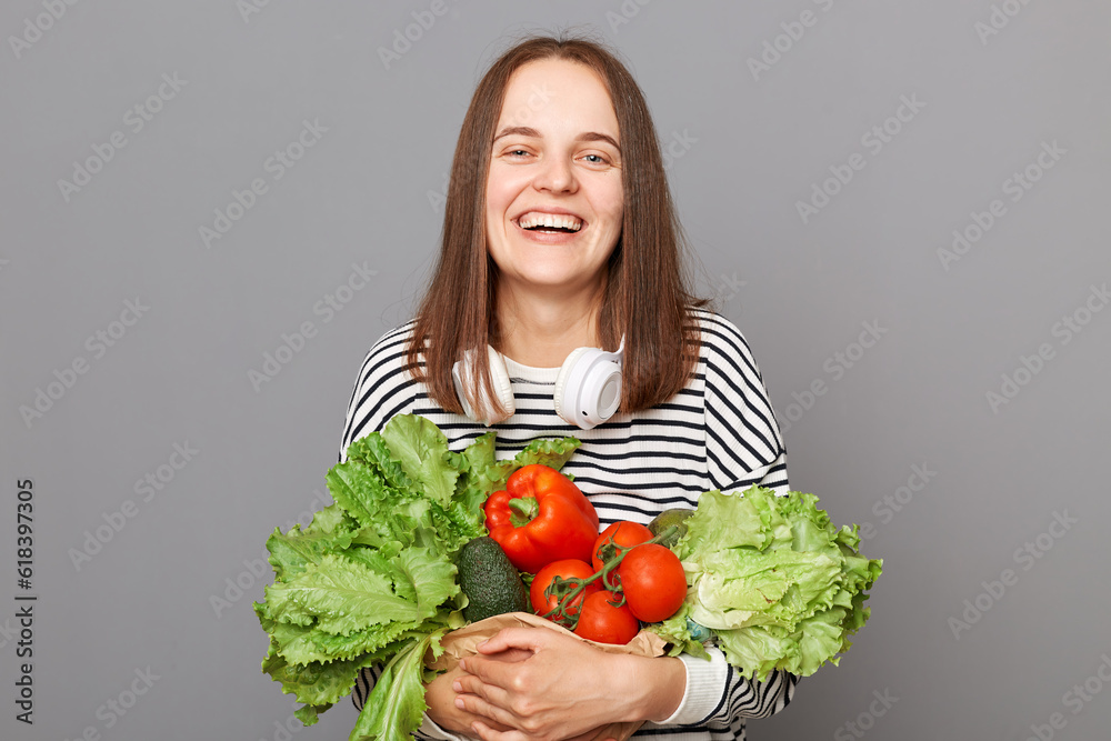 Quality products and vegetables Fresh nutritious for customers. Laughing woman embracing fresh vegetables returns from supermarket wearing striped shirt isolated over gray background Quality products and vegetables Fresh nutritious for customers. Laughing woman embracing fresh vegetables returns from supermarket wearing striped shirt isolated over gray background