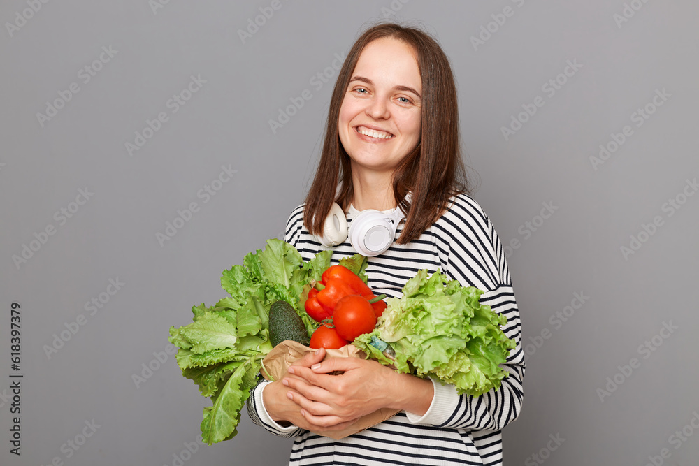 Healthy shopping bag. Fresh food market nutrition. Smiling woman embracing fresh vegetables returns from supermarket wearing striped shirt isolated over gray background Healthy shopping bag. Fresh food market nutrition. Smiling woman embracing fresh vegetables returns from supermarket wearing striped shirt isolated over gray background