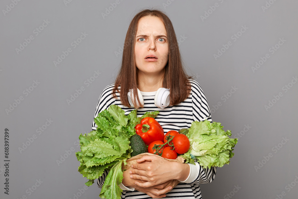 Nourishing food choices at the grocery store. Confused surprised woman embracing fresh vegetables returns from supermarket wearing striped shirt isolated over gray background Nourishing food choices at the grocery store. Confused surprised woman embracing fresh vegetables returns from supermarket wearing striped shirt isolated over gray background