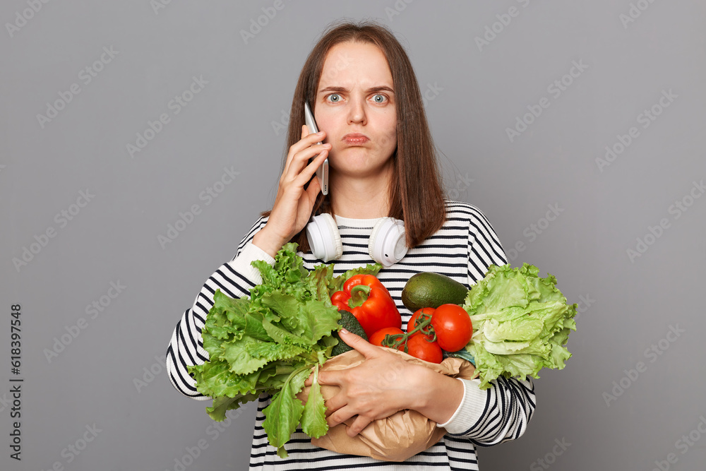 Sad unhappy woman talking on smartphone embracing fresh vegetables returns from supermarket wearing striped shirt isolated over gray background looking at camera with confused look. Sad unhappy woman talking on smartphone embracing fresh vegetables returns from supermarket wearing striped shirt isolated over gray background looking at camera with confused look.
