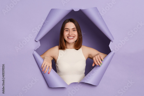 Εκτύπωση καμβά Charming smiling young brunette woman in casual clothing breaking through purple paper hole being in good mood looking with happy face friendly expression