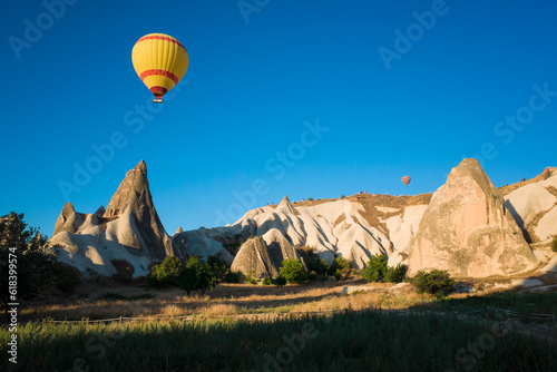 A yellow hot air balloon flies against a clear blue sky over the rock formations of Fairy Chimneys Cappadocia, Goreme National Park, Turkey