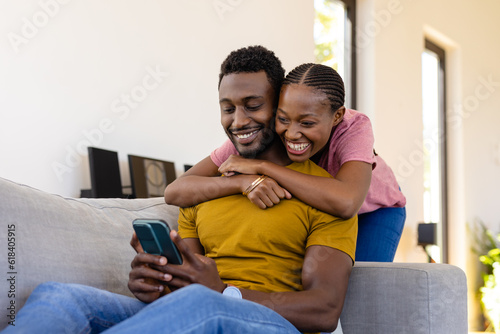 Happy african american couple smiling, embracing and using smartphone on couch at home
