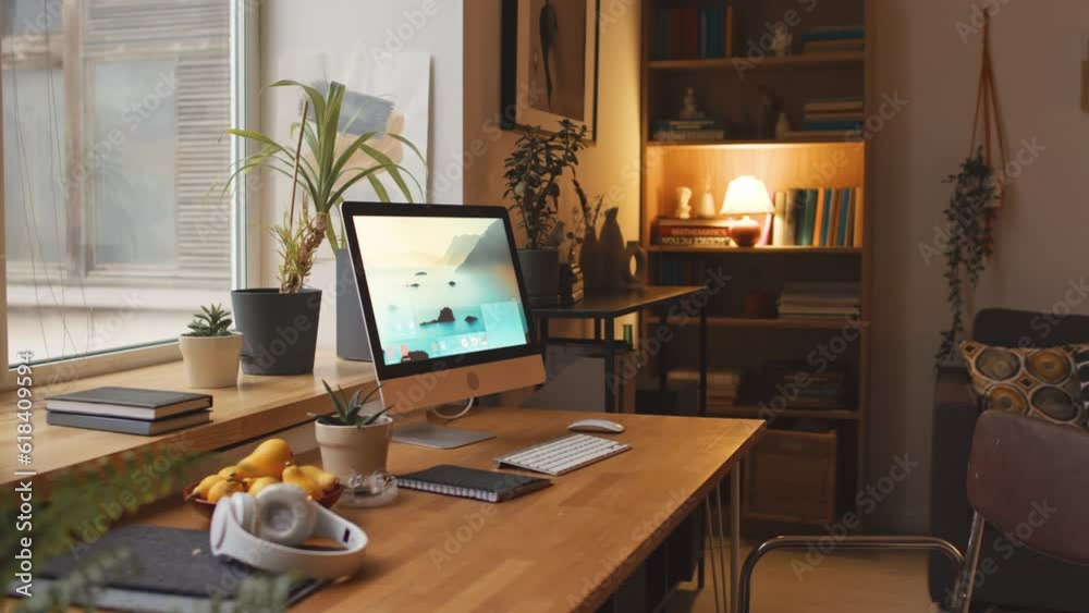 No people shot of working desktop computer on wooden desk by window and old books in bookcase in cozy living room with warm evening light