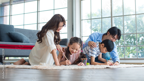 Asian happy cheerful joyful family husband and wife lover couple sitting on carpet floor helping teaching little boy son and girl daughter painting drawing cartoon with color pencils in living room