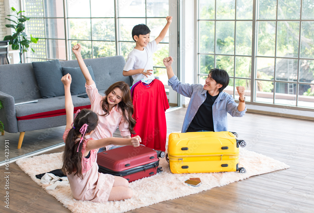 Fototapeta premium Asian little boy and girl helping father and mother preparing packing clothes and stuff into trolley luggages on living room carpet floor for traveling on holiday weekend vacation family road trip