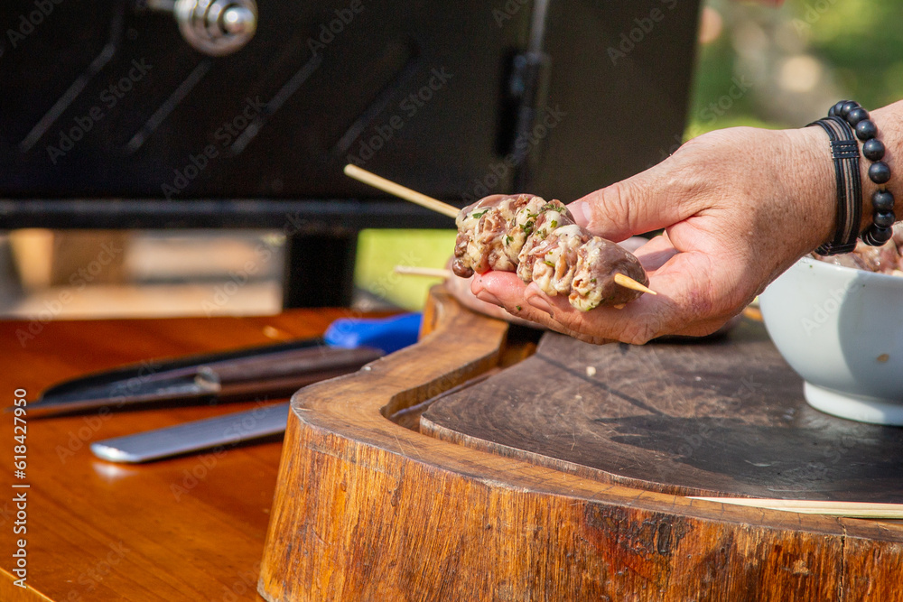 Obraz premium A white man's hand holding a chicken heart stick next to some knives and a white bowl above a rustic wooden board ready to bring it to the barbecue grill for dinner or lunch