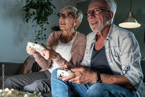 Senior couple sitting on sofa and playing video game on console. They're holding a game pad and challenge each other to win. 
