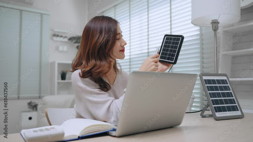 Happy young asian woman holding solar cell panel looking details and record note in book on table in living room at home. Alternative energy and energy saving Happy young asian woman holding solar cell panel looking details and record note in book on table in living room at home. Alternative energy and energy saving