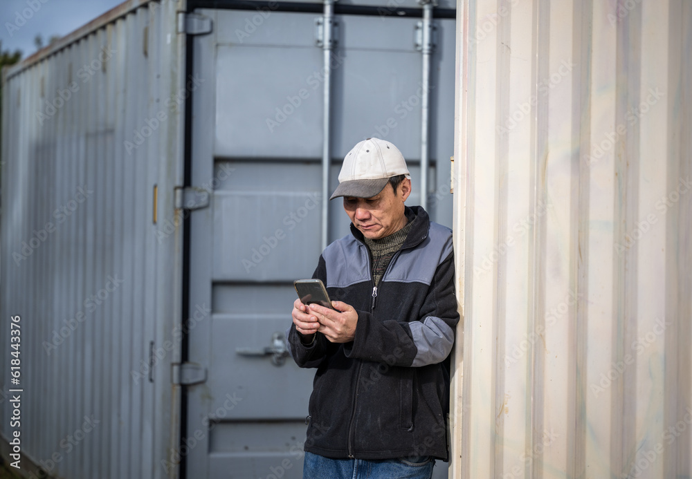 Serious looking worker holding smartphone, standing between containers on construction site. Serious looking worker holding smartphone, standing between containers on construction site.