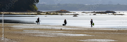 Backlit silhouettes of a male shell fisher and two female shell fishers skirting the shore to enter the water at low tide to shellfish for mussels and clams on a beach. photo