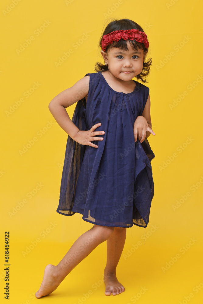 A full length shot of adorable little Asian girl wearing dress standing hands at the waist, isolated on yellow background. A full length shot of adorable little Asian girl wearing dress standing hands at the waist, isolated on yellow background.