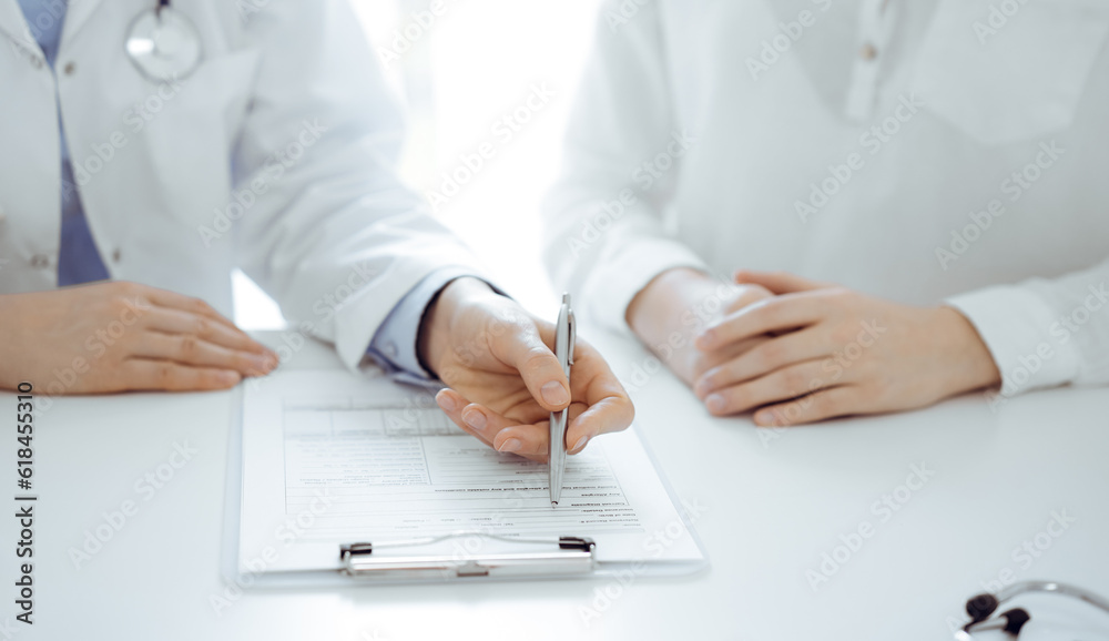 Doctor and patient sitting near of each other at the desk in clinic. The focus is on female physician's hands filling up the medication history record form or checklist, close up. Medicine concept Doctor and patient sitting near of each other at the desk in clinic. The focus is on female physician's hands filling up the medication history record form or checklist, close up. Medicine concept
