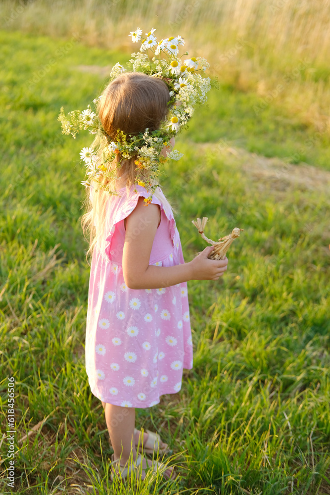 Fototapeta premium small child, happy cool blonde girl 4 years old in pink dress stands in green meadow, kid's head is decorated with cute wreath wildflowers, daisies, grass doll in hands, happy childhood