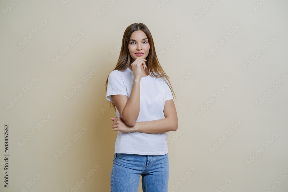 Brunette woman holds chin, wears white t-shirt and jeans, conversing with mates, isolated on brown background Brunette woman holds chin, wears white t-shirt and jeans, conversing with mates, isolated on brown background