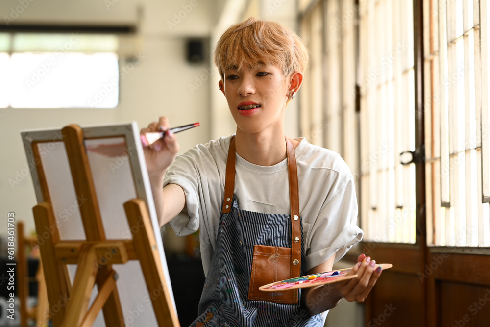 Inspired asian male student holding palette painting picture on canvas in art workshop. Art, education and leisure activity concept Inspired asian male student holding palette painting picture on canvas in art workshop. Art, education and leisure activity concept