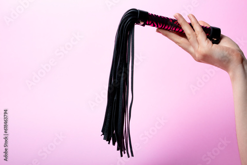 female hand holds a black leather whip with a pink handle on a pink background. Place for text. Sex shop concept. Copy space