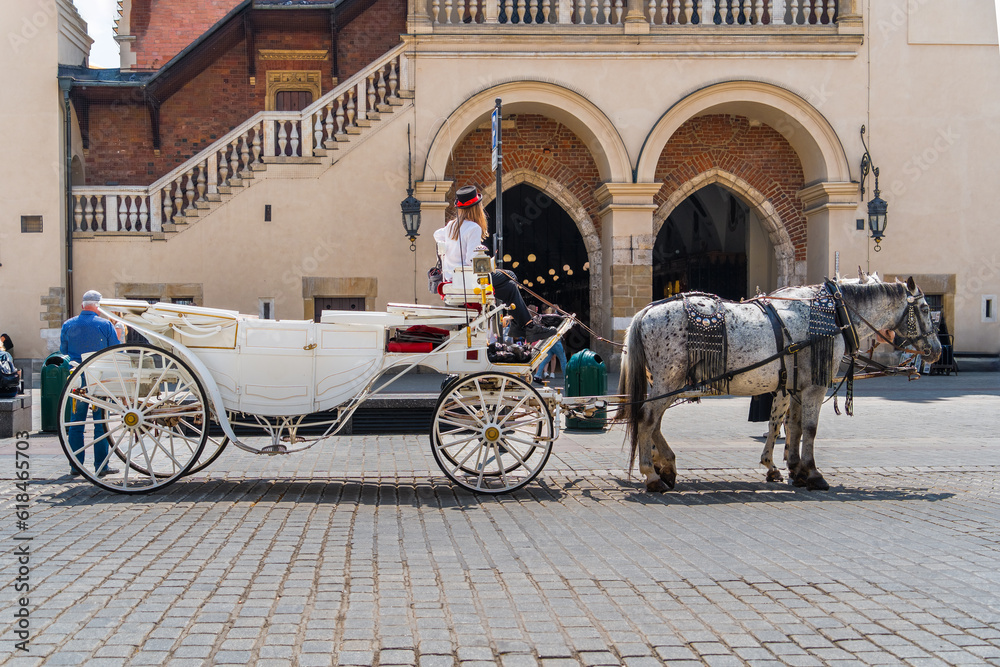 A white horse-drawn carriage and a woman rider wait for tourists on the gray cobbled street of the old town on a sunny day at the entrance to the old main market of Krakow. Horse transport in Europe