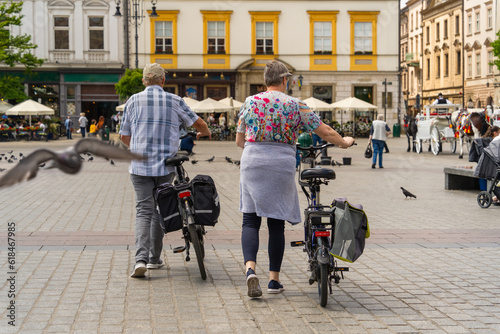 Authentic elderly couple with electric bikes in the city square on a sunny day against the background of tourists, pigeons, horse-drawn carriages, street cafe umbrellas, clean urban transport photo