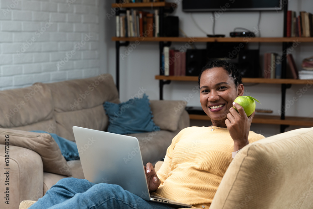 African American woman reading online PDF book and eating apple in living room. Healthy life and enjoyment at home in free time.