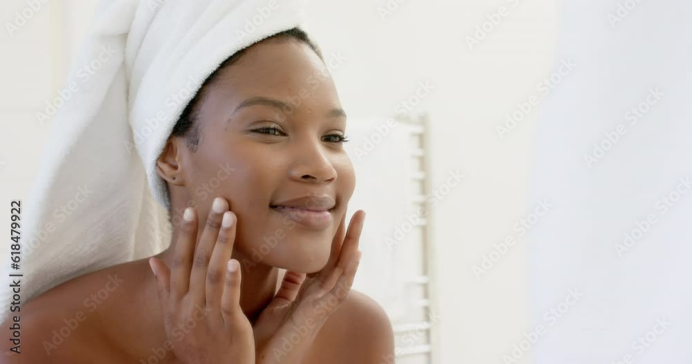Happy biracial woman with towel on head looking in mirror touching her face in bathroom, slow motion Happy biracial woman with towel on head looking in mirror touching her face in bathroom, slow motion
