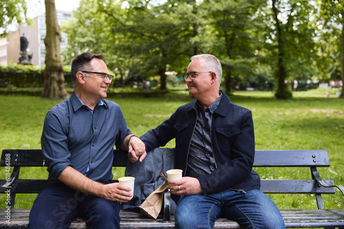 Same sex male couple sitting on park bench chatting drinking coffee and holding hands