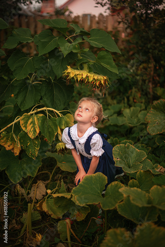 A little blonde girl in a blue rustic dress stands in front of a large sunflower flower
