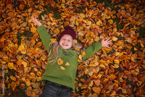 A little beautiful blonde girl in a burgundy hat and a green hoodie lies in red autumn leaves, top view