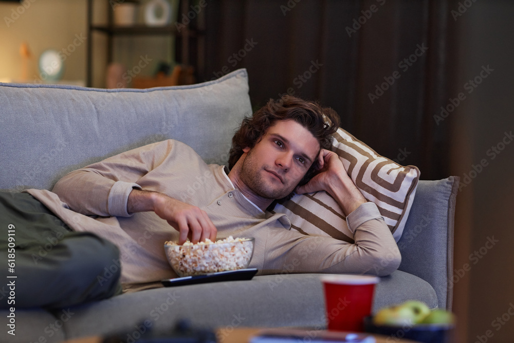 Portrait of young man watching Tv at home and eating popcorn while lying on couch, copy space