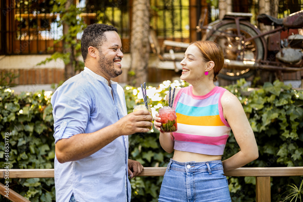 Fototapeta premium A multi-ethnic couple happily drinks mojitos on the terrace of a bar. African man and Caucasian woman look at each other while drinking alcoholic cocktails. Interracial love.