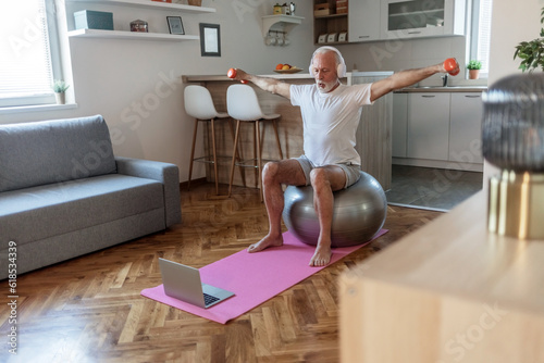 Positive senior man exercising with dumbbells  sitting on fitball and listening to music in headphones indoors.