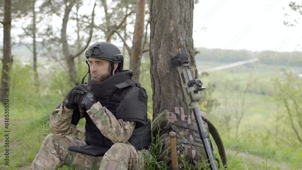 Mine clearance. Soldier with mine detector and backpack sits exhausted by a tree after heavy demining. Demining process Mine clearance. Soldier with mine detector and backpack sits exhausted by a tree after heavy demining. Demining process