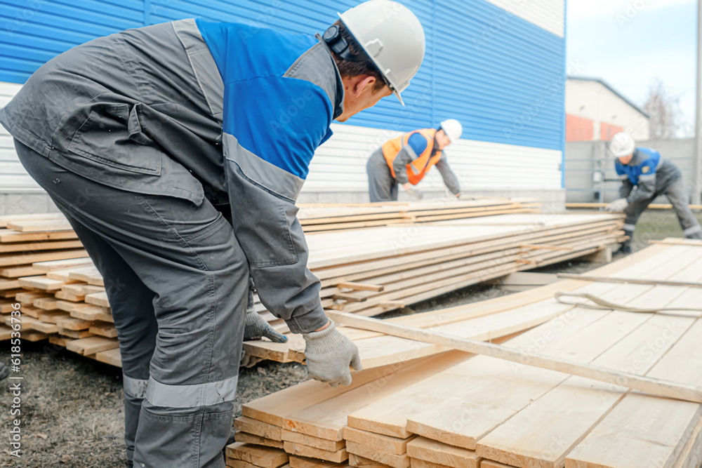Workers in hard hats and overalls stack wooden boards outside on street. Procurement and sale of building materials. Industrial background. Workers in hard hats and overalls stack wooden boards outside on street. Procurement and sale of building materials. Industrial background.