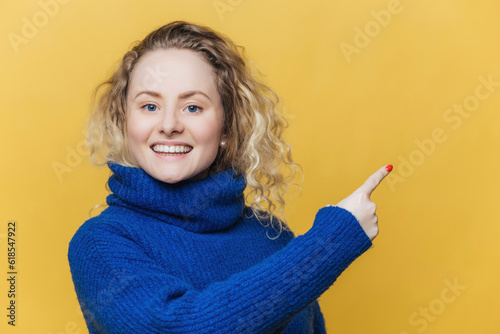 Happy blonde woman in blue sweater advertises with joy, pointing at blank space on yellow wall. photo